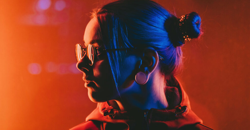 Millennial pretty girl with unusual hairstyle near glowing red neon of city at night. Dyed blue hair in braids. Mysterious hipster teenager in glasses. Reflection of light.